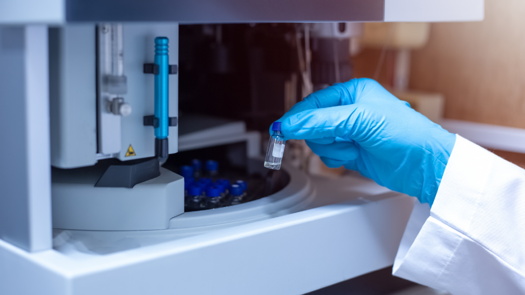 A gloved hand holds a small vial near an analytical laboratory instrument containing several other vials.